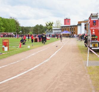 Zdjęcie przedstawia boisko GOKiS podczas zawodów, widać uczestników i sędziów na płycie boiska oraz widownię na trybunach. Po prawej stronie stoi wóz strażacki OSP Ruda.