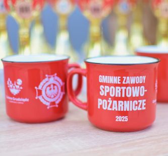 Zdjęcie przedstawia kubki okolicznościowe przygotowane na zawody oraz na drugim planie puchary. 